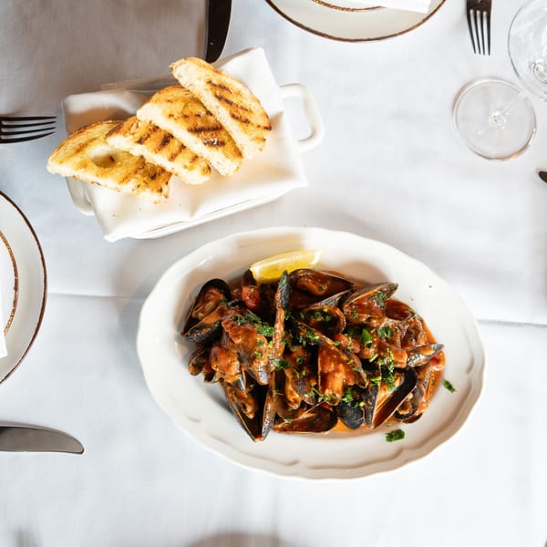 Mussels in red sauce with parsley and a lemon wedge, served with grilled bread on a white tablecloth.