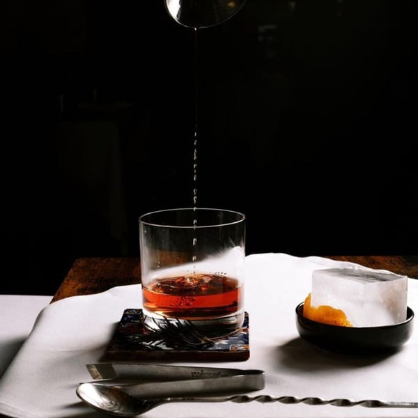 A dark cocktail is poured into a glass, next to a large ice cube with orange peel.