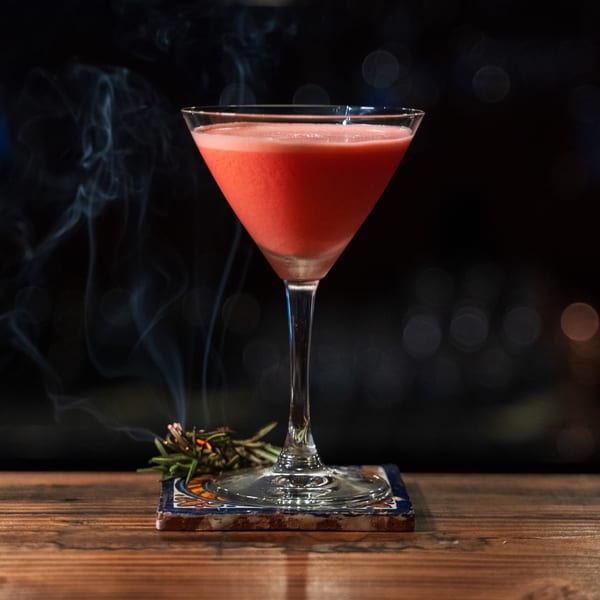 Red cocktail in a martini glass on a patterned coaster with rosemary and smoke on a wooden table.