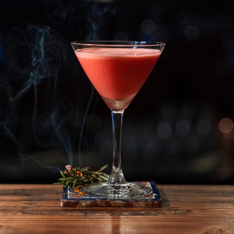 Red cocktail in a martini glass on a patterned coaster with rosemary and smoke on a wooden table.