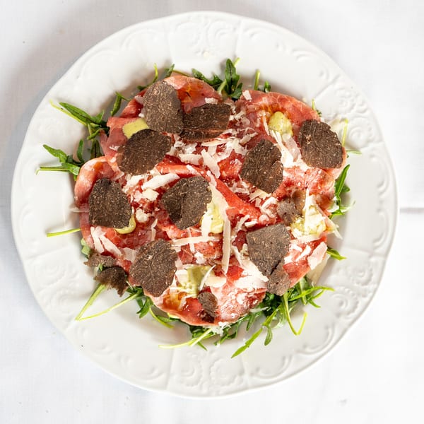 Antica Sicilia's carpaccio with shaved truffles, parmesan, and arugula on a white plate.