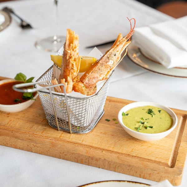 Fried shrimp in a metal basket with lemon, red sauce, and green sauce on a wooden board.