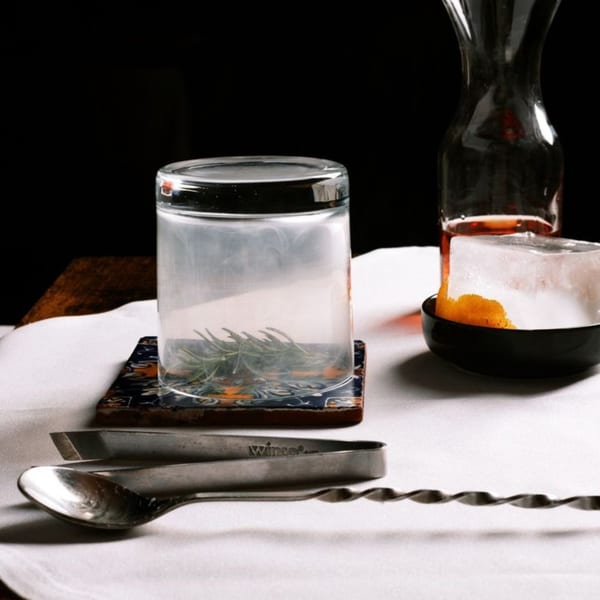 A glass with rosemary, a carafe, ice with orange peel, and bar tools on a white tablecloth.