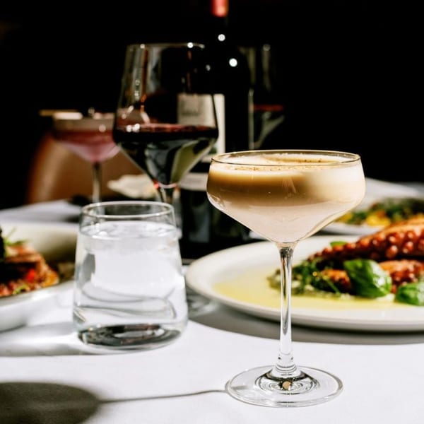 A creamy cocktail, red wine, water, and Italian dishes on a white tablecloth at Antica Sicilia.