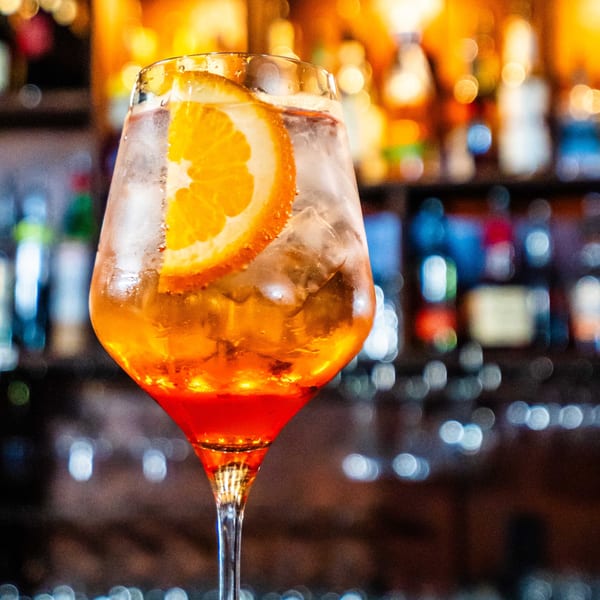 A vibrant orange Aperol Spritz cocktail with ice and an orange slice, against a blurred bar background.