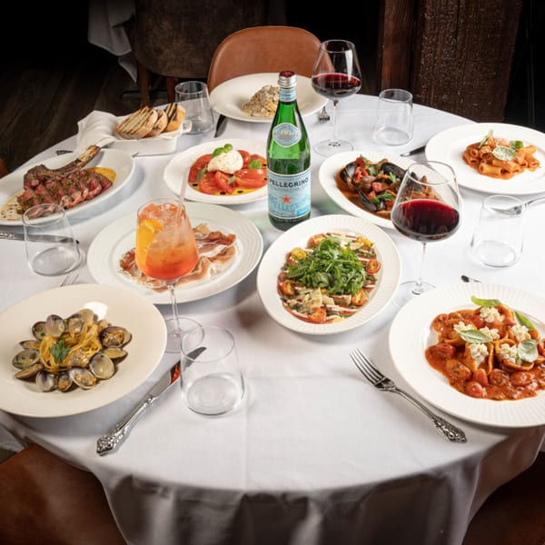 A white-clothed table at Antica Sicilia laden with various Italian dishes, wine, and a sparkling water bottle.