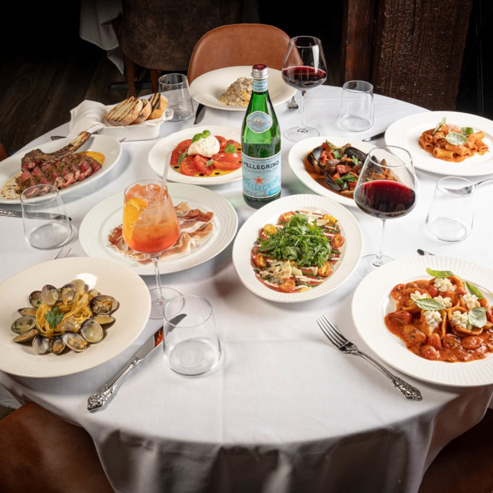 A white-clothed table at Antica Sicilia laden with various Italian dishes, wine, and a sparkling water bottle.